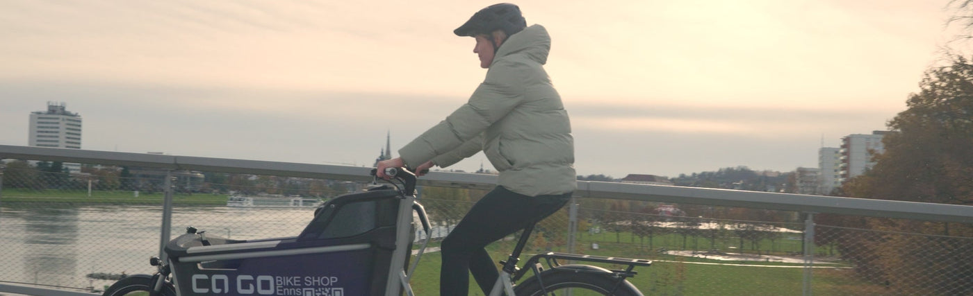 Mutter mit Kind fährt mit Cago FS200 Cargo Bike und Eightpins NGS2 Cargo Sattelstütze über eine Brücke mit der Donau und der Stadt Linz im Hintergrund.